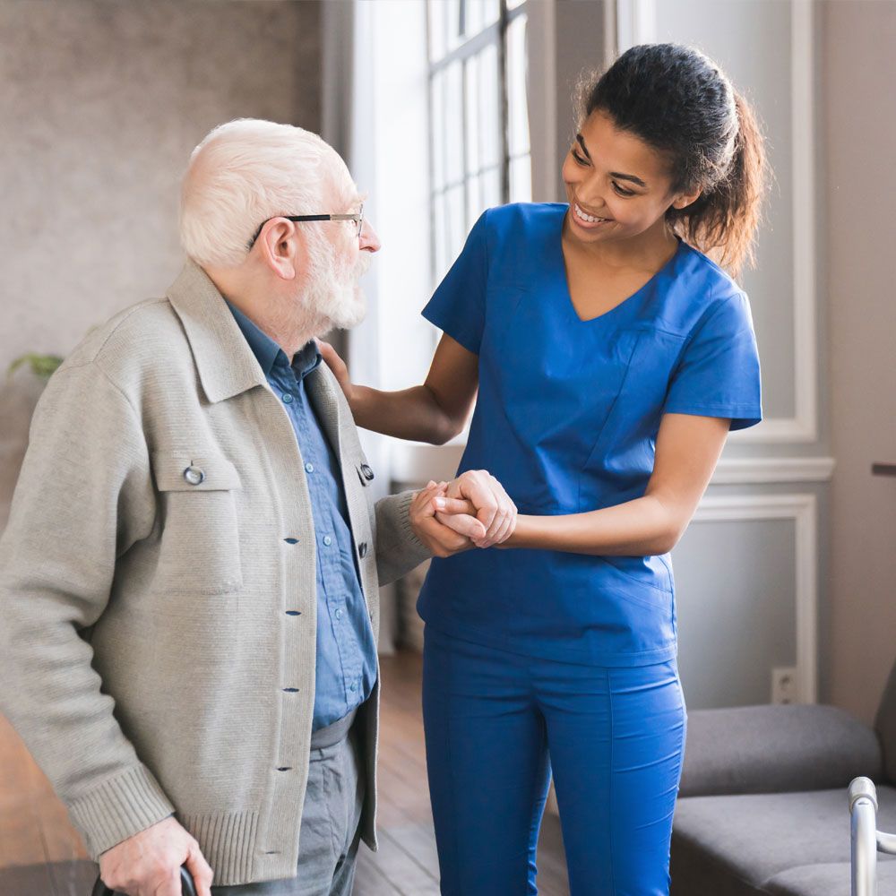 Eine Pflegekraft in blauem Kittel hilft einem älteren Patienten beim Gehen.