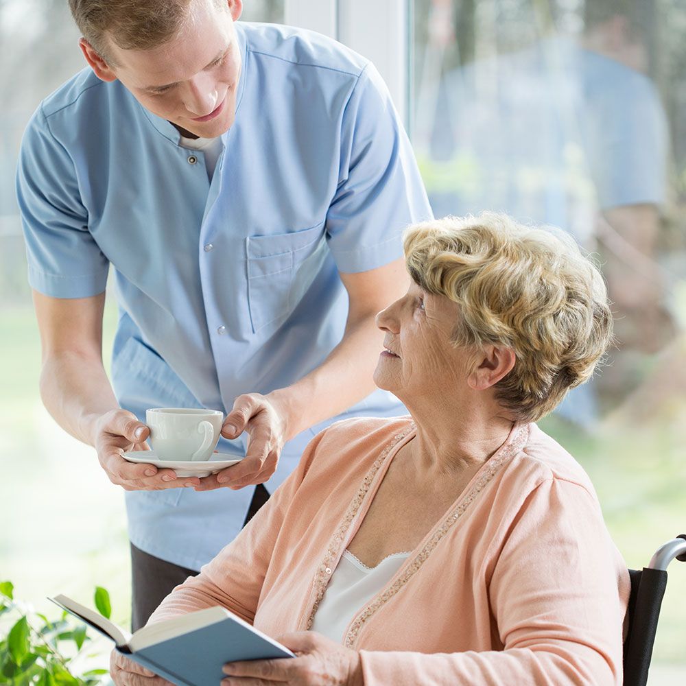 Eine Jungeperson bietet einer älteren Person, die ein Buch liest, eine Tasse mit einem Heißgetränk an